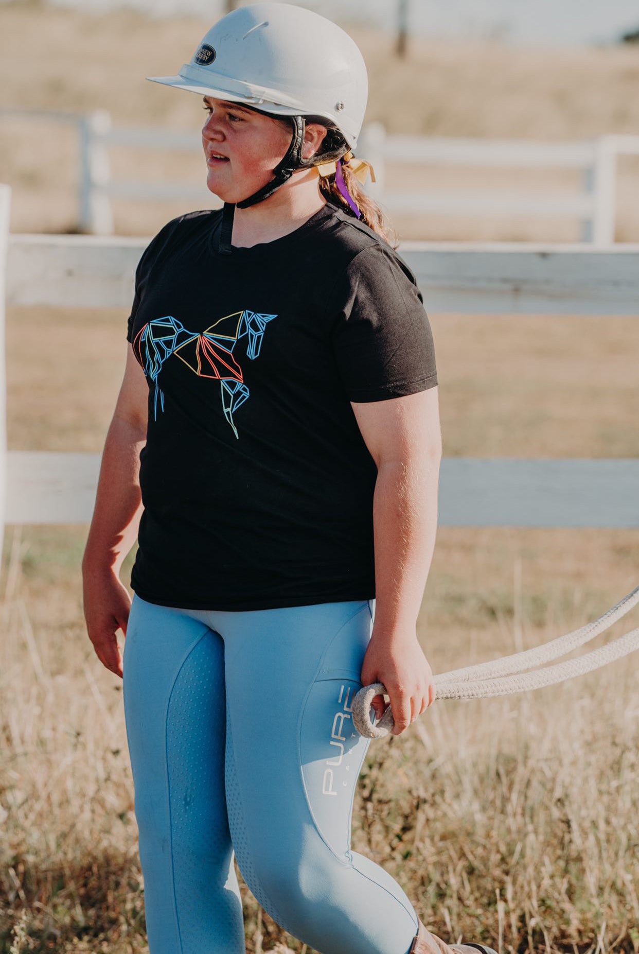 A young person with long hair stands outdoors wearing a white helmet, black Pure Canter Youth PURE Emblem Shirt – a true wardrobe staple – and light blue pants. They hold a rope in their left hand. In the background, there is a white fence and a grassy field.