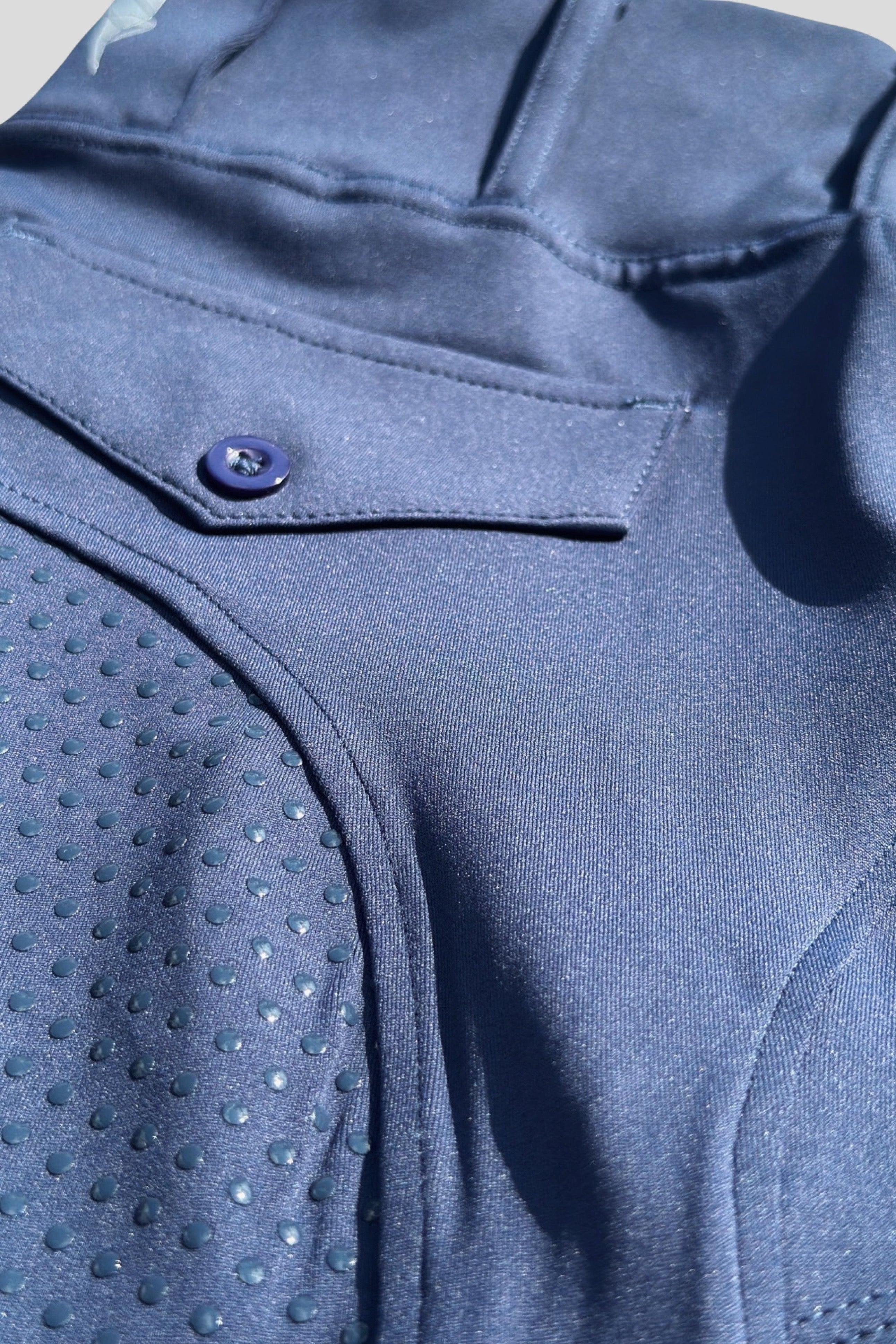 Close-up image of a section of a dark blue garment, likely V2 Fusion Riding Tights - Navy by Pure Canter. The fabric features a smooth texture and a dotted, textured pattern on one part. A visible button and pocket detail are also present, as well as stitching lines. The image is well-lit, showcasing equestrian fashion at its finest.