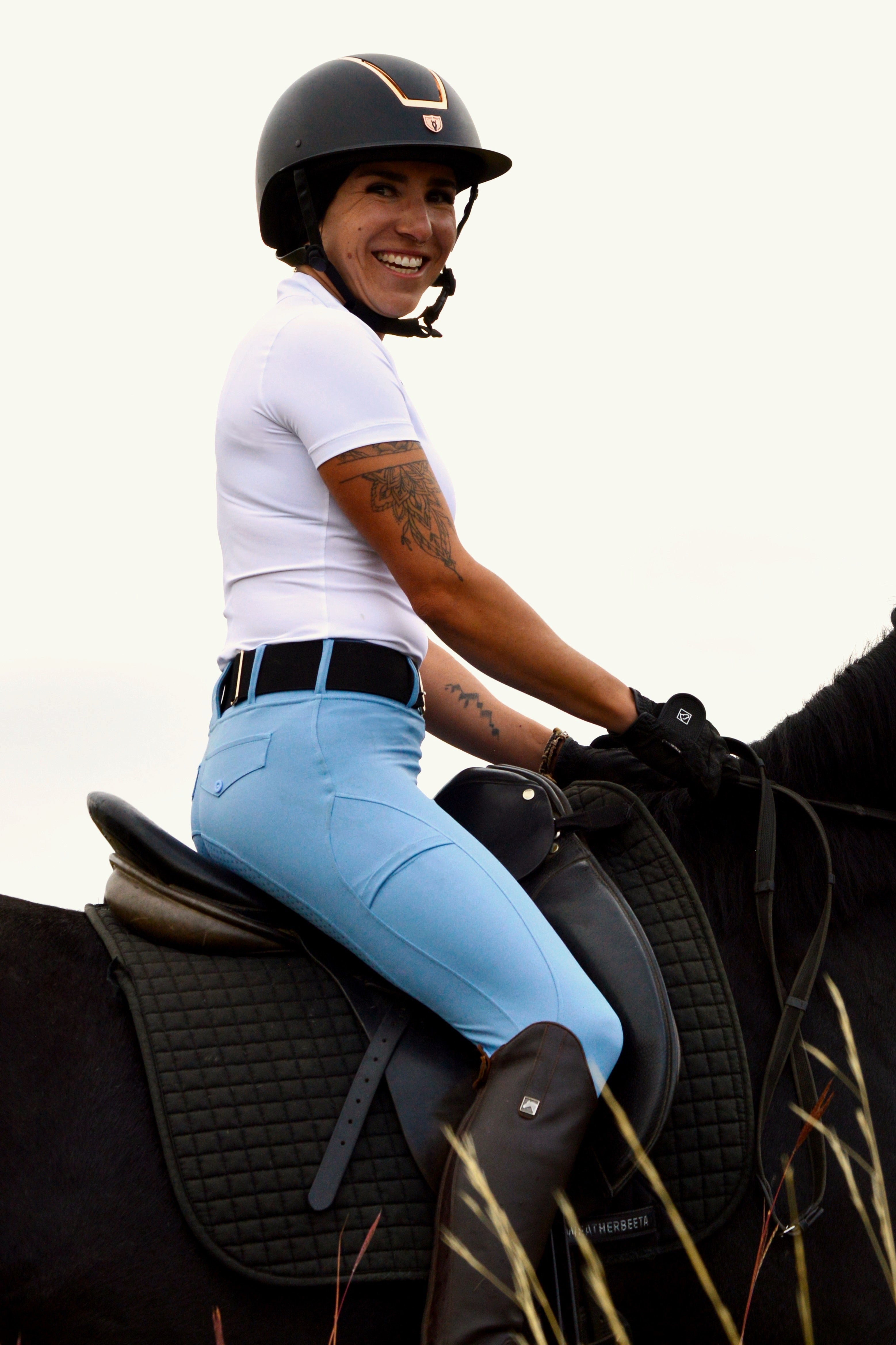 A person wearing a helmet, white shirt, and Pure Canter V1 Fusion Riding Tights - Baby Blue with a high waistband smiles while sitting on a black horse. The person has visible tattoos on their arm and wears brown riding boots. The image focuses on the rider, with the horse's saddle and part of its body in view.