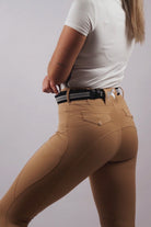 A person stands with their back to the camera, wearing a white short-sleeved shirt tucked into Pure Canter V1 Fusion Riding Tights - Tan. They have a black belt with a silver buckle, and their light brown hair is partially visible. The focus is on the upper body and waist area, showcasing equestrian fashion.