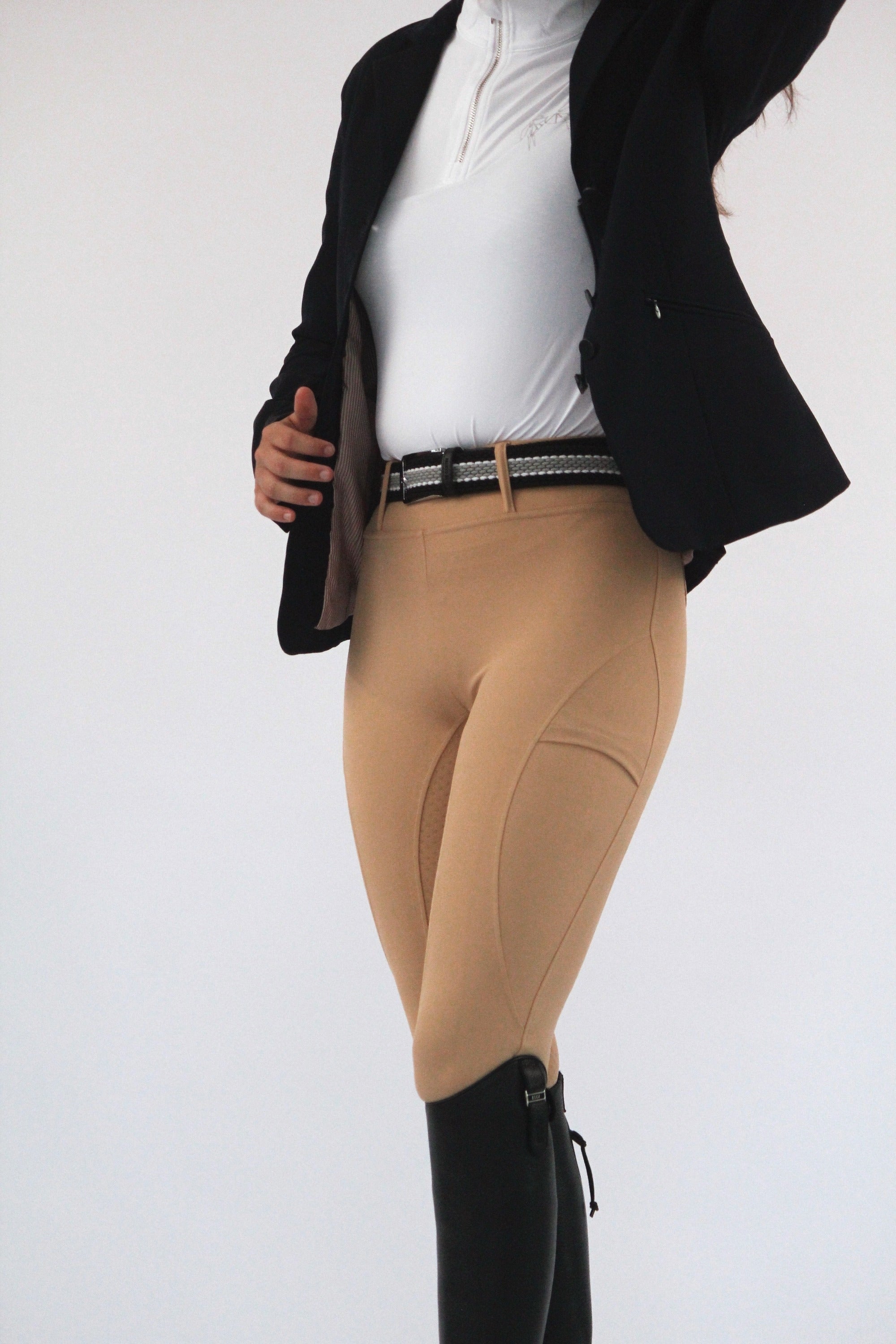 A person wearing Pure Canter's V1 Fusion Riding Tights - Tan, a white shirt, a dark blazer, and black riding boots is shown from the shoulders down. The individual is adjusting their blazer in this elegant equestrian fashion ensemble against a white background.