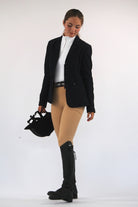 A person stands against a plain white background, dressed in equestrian attire that showcases the best of equestrian fashion. They wear a black jacket, white shirt, V1 Fusion Riding Tights - Tan by Pure Canter made from performance fabric, and black knee-high boots. Holding a black riding helmet in their left hand, they look down slightly.