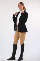 A person stands against a plain white background wearing an equestrian outfit, showcasing equestrian fashion with a black blazer, white shirt, V1 Fusion Riding Tights - Tan from Pure Canter, and black riding boots. They have their hair pulled back and gaze slightly downward while adjusting their blazer.