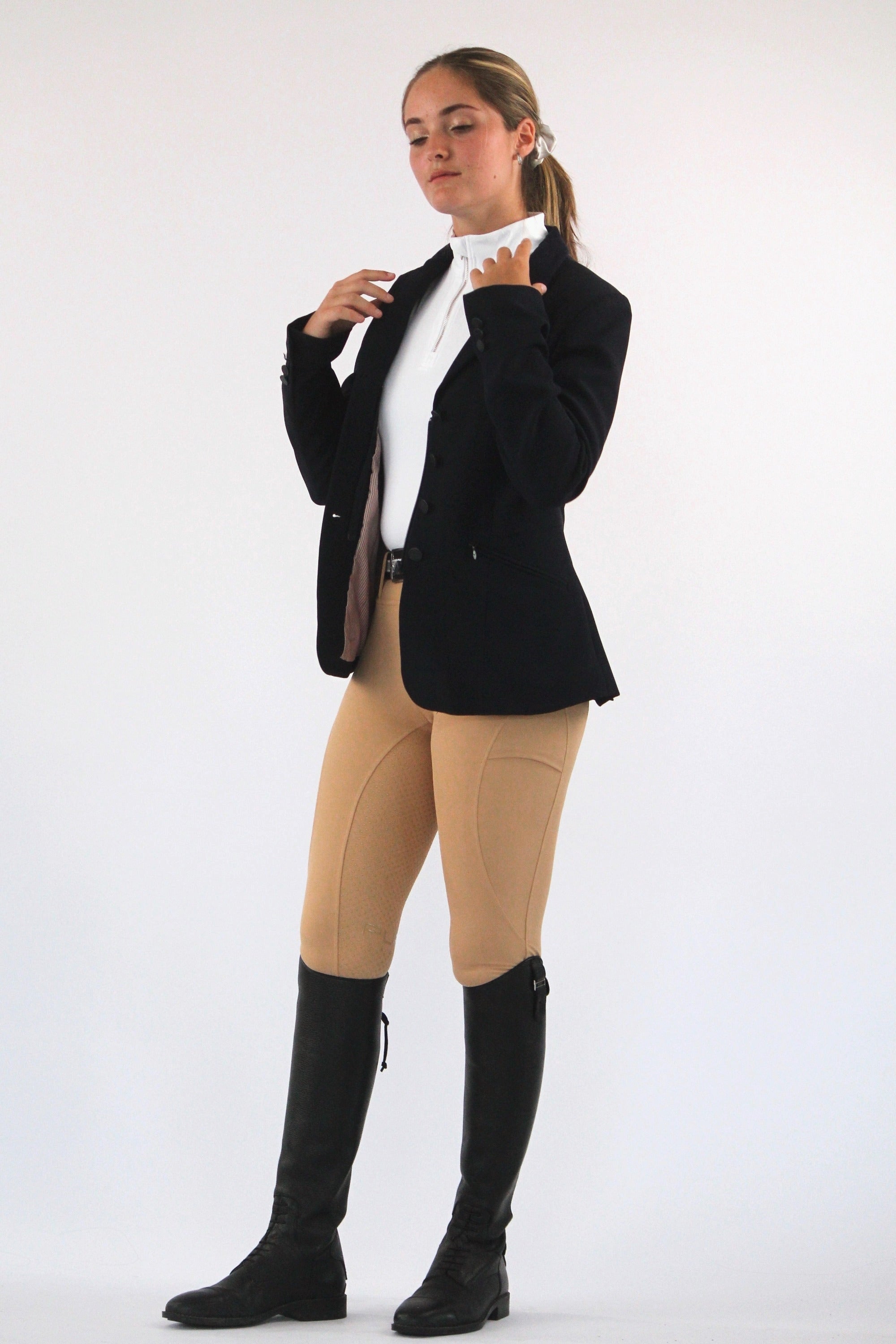 A person stands against a plain white background wearing an equestrian outfit, showcasing equestrian fashion with a black blazer, white shirt, V1 Fusion Riding Tights - Tan from Pure Canter, and black riding boots. They have their hair pulled back and gaze slightly downward while adjusting their blazer.