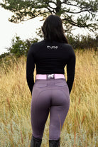 A person with long hair wearing a black long-sleeved shirt, Pure Canter V1 Fusion Riding Tights - Plum with silicone grip, and a pink belt stands facing away in a grassy field. A tree is visible in the background. The shirt has the word "PURE" written on the back.