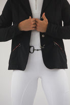 A person is wearing a tailored black blazer over a white zip-up top and Pure Canter Competition Tights - White. The person's hands grasp the edges of the blazer. The outfit includes a fashion accessory resembling a horse bit and features silicone grip details. The background is plain white.