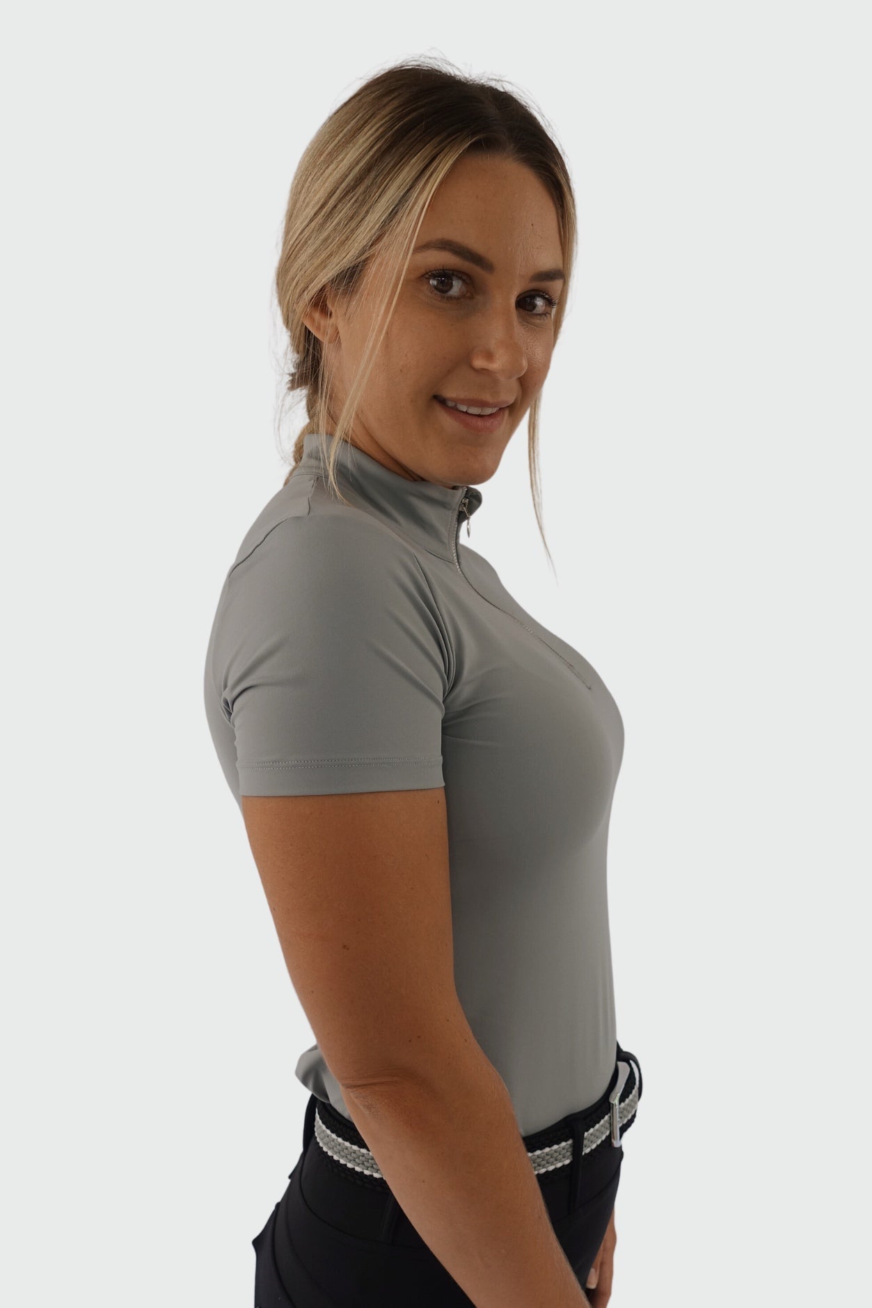 A person with long blond hair is standing in a side profile, turned slightly towards the camera, smiling. They are wearing a gray Pure Canter V1 Motion Short Sleeve and dark pants with a white braided belt against a plain white background.