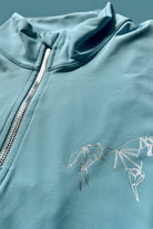 Close-up of a light blue zip-up V1 Motion Top Short - Ocean Blue with a raised collar by Pure Canter. The breathable jacket features a white geometric horse design on the chest. The silver zipper extends from the bottom to the top of the collar. The background is a similar shade of light blue.