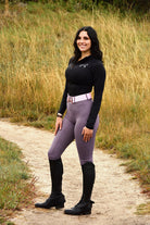 A person with long dark hair stands smiling on a dirt path through tall grass. They are wearing a black long-sleeved top, V1 Fusion Riding Tights - Plum by Pure Canter, a pink belt, and black riding boots. Their hands are relaxed by their sides with one resting on their hip.