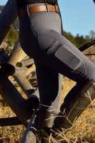 A person dressed in Pure Canter V2 Fusion Riding Tights - Dark Grey with reinforced knee patches, tall brown riding boots, and a brown leather belt leans against a wooden fence in a field. The background features dry grass, trees, and a clear blue sky, embodying classic equestrian fashion.