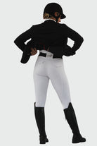 A person in equestrian attire stands with their back to the camera. They are wearing a black riding helmet, black jacket, Pure Canter Competition Tights - White, and black riding boots. The person appears to be adjusting their jacket made from performance fabric. The background is plain white.
