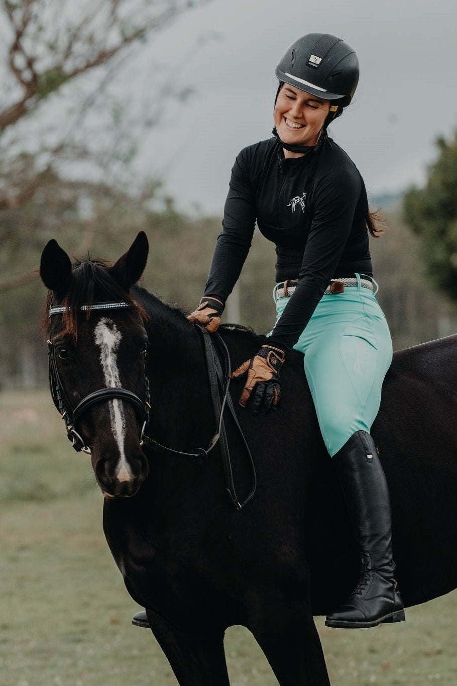 A person in a black helmet and black long-sleeve shirt rides a dark horse. The rider wears Pure Canter's V1 Fusion Riding Tights - Aqua with silicone grip, completed by black boots and gloves. They are smiling, looking down at the horse. The background reveals a blurred outdoor setting with trees and greenery.