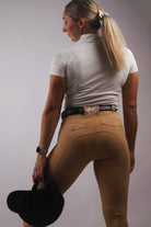 A woman stands with her back to the camera, holding a black riding helmet in her right hand. Clad in a white short-sleeve shirt and Pure Canter V1 Fusion Riding Tights - Tan, she accessorizes with a black belt and an Apple Watch on her left wrist. Her ponytail of blonde hair ties the equestrian fashion look together.