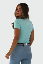 A person with long hair is standing and looking over their shoulder towards the camera. They are wearing the Pure Canter V1 Motion Top Short - Ocean Blue made from breathable fabric and matching blue pants with a silver belt and horse emblem on the back pocket.