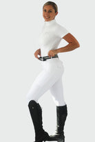 A person is posing and standing sideways, wearing a white short-sleeve riding shirt tucked into white Pure Canter Competition Tights made from performance fabric. The person has a black belt, black knee-high riding boots, and is smiling. The background is plain white.