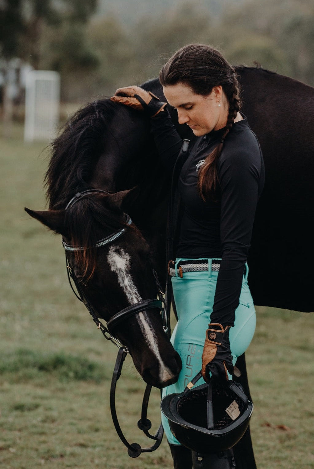 A woman wearing equestrian gear, including a black shirt and Pure Canter Fusion Tights - Aqua with silicone grip features, gently touches and stands next to a black horse with a white blaze on its face. She holds a riding helmet in her other hand, and they are outdoors in a grassy area.
