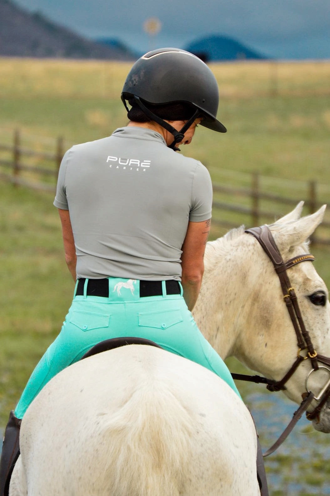 A person wearing a helmet and a gray shirt with "Pure Canter" written on the back is riding a white horse. With fields and a wooden fence in the background, the rider faces away from the camera, showcasing their V1 Motion Short Sleeve - Grey designed for ultimate comfort.