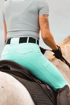 A person riding a horse, seen from the side. The rider is wearing a light gray shirt, Pure Canter V1 Fusion Riding Tights - Aqua, black gloves, and a black belt. The horse has a light-colored coat and dark saddle. The rider's arm reveals a tattoo.