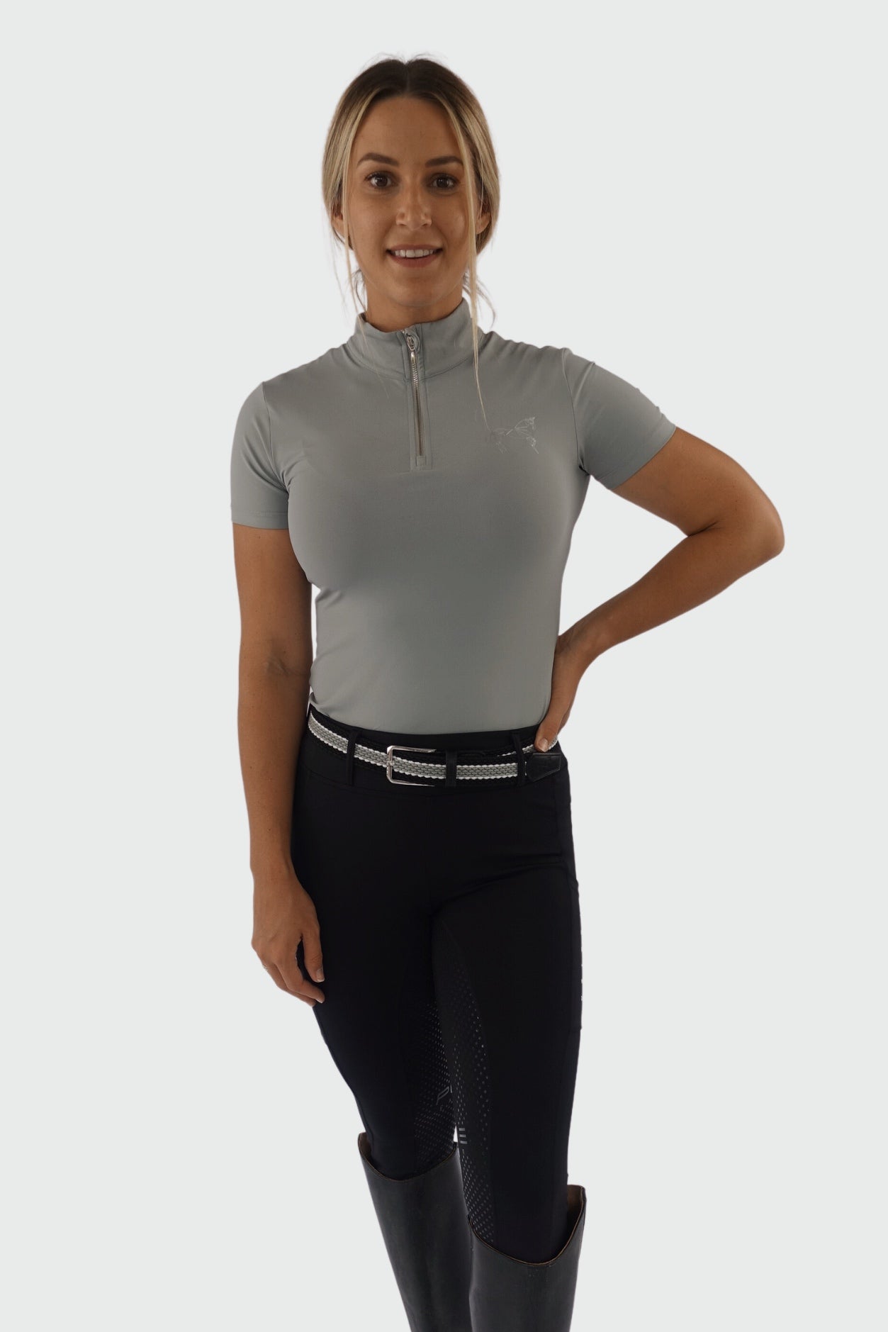 A woman with blond hair, tied back, wearing a short-sleeved, light gray zip-up top made of premium base layers called the V1 Motion Short Sleeve - Grey by Pure Canter, black riding pants, and black knee-high boots stands against a plain background. One hand rests on her hip while the other hangs by her side. She is smiling at the camera.