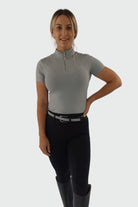 A woman with blond hair, tied back, wearing a short-sleeved, light gray zip-up top made of premium base layers called the V1 Motion Short Sleeve - Grey by Pure Canter, black riding pants, and black knee-high boots stands against a plain background. One hand rests on her hip while the other hangs by her side. She is smiling at the camera.