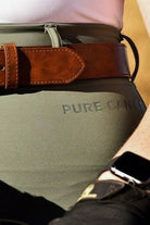 Close-up photo of a person wearing the Pure Canter V2 Fusion Riding Tights - Sage with the partially visible text "PURE GANG," reminiscent of equestrian fashion, and a brown leather belt with a large buckle. The person is also wearing a smartwatch on their right wrist.