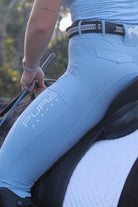 A person wearing light blue V1 Fusion Riding Tights - Baby Blue by Pure Canter with a high waistband and a matching shirt is seated on a horse. The pants, featuring the words "PURE" and "CANTER" printed on the thigh, have a silicone grip. The person holds the reins and uses a grey smartwatch. The saddle has a white quilted cover.