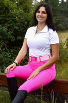 A woman with dark hair sits on a wooden bench outdoors, surrounded by greenery. She is smiling and wearing a white polo shirt, pink belt, and bright pink V2 Fusion Riding Tights - Hot Pink by Pure Canter, complemented by black knee-high boots. Her ensemble perfectly encapsulates modern equestrian fashion.