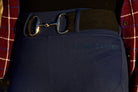 Close-up of a person wearing blue Fusion Tights G2 - Navy and a belt featuring a horse bit buckle. The waistband of the pants has "PURE CANTER" printed on it. The person is also wearing a red plaid shirt, showcasing an impeccable touch of equestrian fashion.