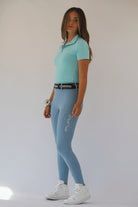 A person stands against a plain background, wearing a light blue polo shirt and matching Pure Canter V1 Fusion Riding Tights - Baby Blue with "PURE" written on the leg. They have long brown hair and are wearing white high-top sneakers. A high waistband black belt and a scrunchie adorn their waist and wrist, respectively.