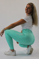 A person with long, blonde hair wearing a white t-shirt and V1 Fusion Riding Tights - Aqua by Pure Canter is squatting on one leg while smiling at the camera. They are also wearing white sneakers and a watch on their left wrist, set against a plain white background.