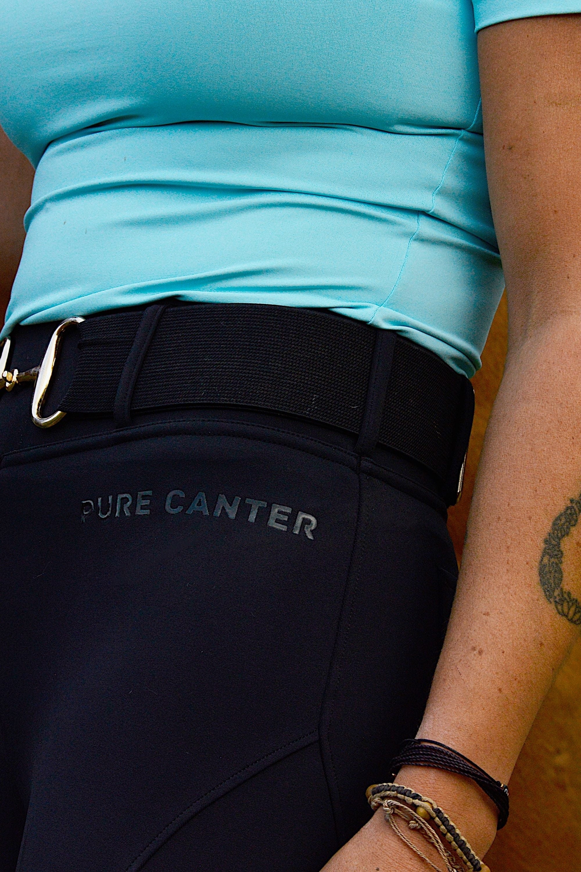 Close-up of a person wearing V2 Fusion Riding Tights - Black by Pure Canter with "Pure Canter" written on the side and featuring a silicone grip. The person is also wearing a light blue shirt, a black belt with a silver buckle, and multiple bracelets on one wrist. A tattoo is visible on their forearm.