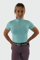 A woman is standing and posing against a plain background, wearing the V1 Motion Top Short - Ocean Blue by Pure Canter and purple riding pants with a black and silver belt. She has her hands placed on her hips and is looking forward with a neutral expression.