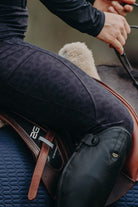 A person wearing Pure Canter Fusion Tights - Black Leopard and black riding boots is seated on a horse saddle. They are holding the reins, with part of the saddle and saddle pad visible in the image. The focus is on the person's lower torso and upper leg area, showcasing the performance fabric of their gear.