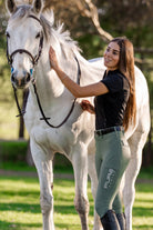 A woman with long brown hair, wearing a black shirt and green Pure Canter Fusion Tights - Sage, stands beside a white horse. She is gently petting the horse's neck with one hand while holding its reins with the other. The scene is set outdoors in a grassy area with trees in the background.