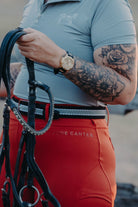 A person with tattooed arms wearing a blue shirt and bright red pants holds horse reins. The individual sports a gold watch on their left wrist. Blending equestrian fashion, the shirt features a subtle horse design while the pants, V2 Fusion Riding Tights - Outback from Pure Canter, are made of performance fabric and have "PURE CANTER" written on the hip.