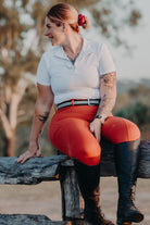 A person with tattoos on both arms sits on a wooden fence outdoors. They are dressed in a white polo shirt and V2 Fusion Riding Tights - Outback by Pure Canter, paired with black knee-high riding boots. The individual has light brown or blonde hair tied back with a red scrunchie, embodying equestrian fashion.