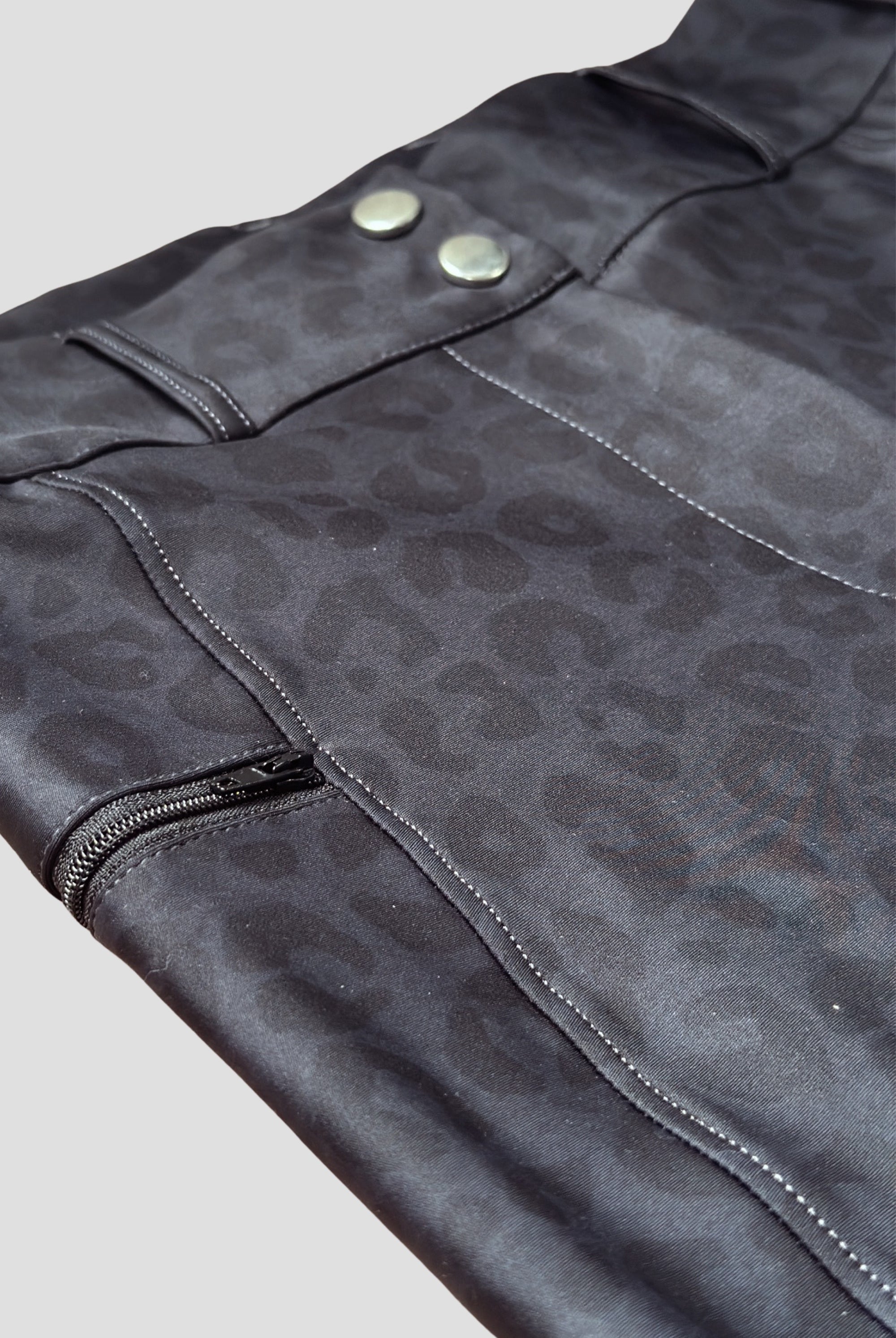 Close-up of a black leather jacket with a subtle leopard print pattern, resembling chic "The Breeches - Black Leopard." The jacket features metal snap buttons, visible white stitching, and a partially open zipper on the left side. The background is plain and light-colored.