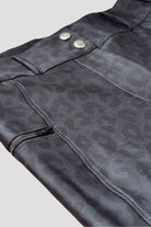 Close-up of a black leather jacket with a subtle leopard print pattern, resembling chic "The Breeches - Black Leopard." The jacket features metal snap buttons, visible white stitching, and a partially open zipper on the left side. The background is plain and light-colored.