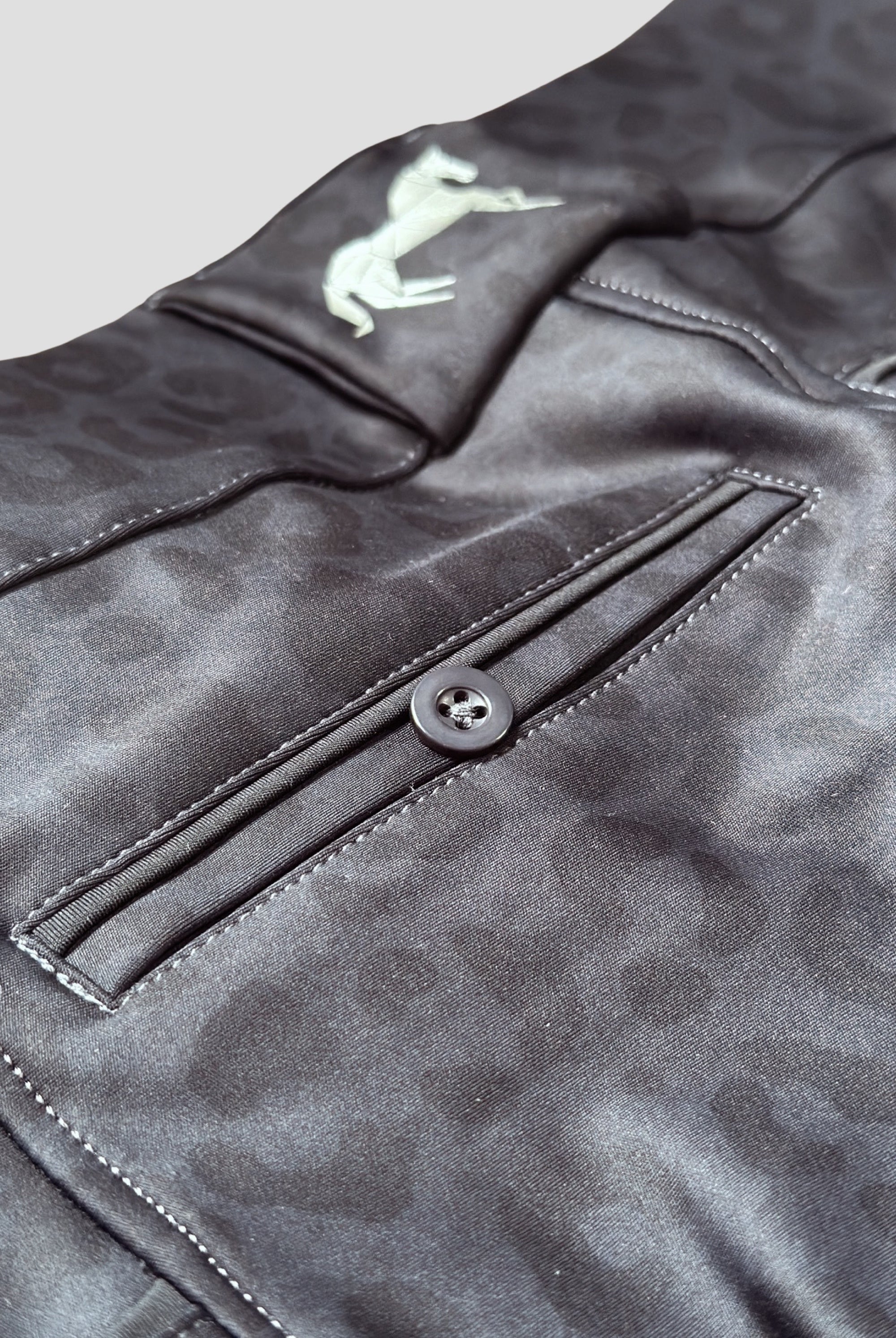 Close-up of dark gray Pure Canter The Breeches - Black Leopard with a subtle leopard print pattern. The image shows a buttoned back pocket, detailed stitching, and an embroidered white logo featuring a horse and rider. Made from performance fabric with a Silicone Seat for added grip, the texture and design details are clearly visible.