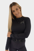 A woman with long blonde hair is smiling at the camera. She is wearing a black, long-sleeved athletic top with a small logo on the chest and black leggings from the Pure Canter Motion Top Long - Black collection. Equipped with premium base layers and a belt with metallic fastenings, she stands confidently against a plain white background.