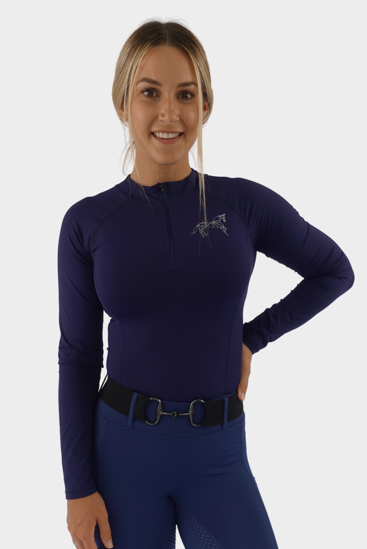 A woman with blonde hair is smiling at the camera. She is wearing a long-sleeve, navy athletic top with a partial zipper and a Pure Canter logo on the chest, along with matching purple athletic pants. Her premium base layers feature quick drying technology. Her right hand is on her hip.