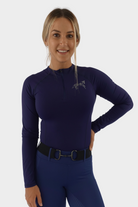 A woman with blonde hair is smiling at the camera. She is wearing a long-sleeve, navy athletic top with a partial zipper and a Pure Canter logo on the chest, along with matching purple athletic pants. Her premium base layers feature quick drying technology. Her right hand is on her hip.