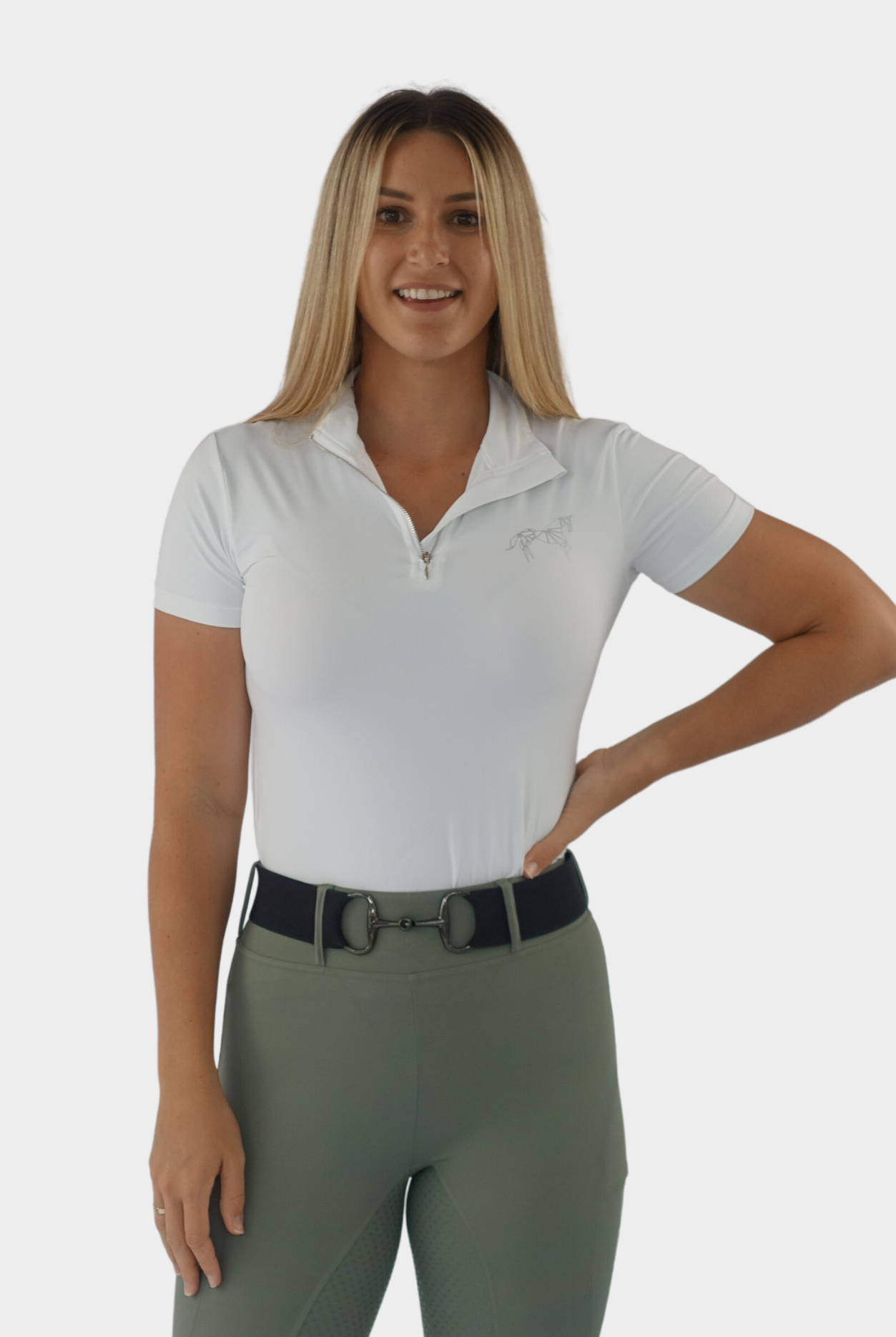 A woman with long blonde hair stands smiling against a plain background. She is wearing a white short-sleeved zip-up shirt and olive green pants with a belt, perfect for rider wear. Her right arm is bent at the elbow with her hand resting on her hip, and her left arm is relaxed by her side. She is sporting the Motion Top Short - White from Pure Canter.