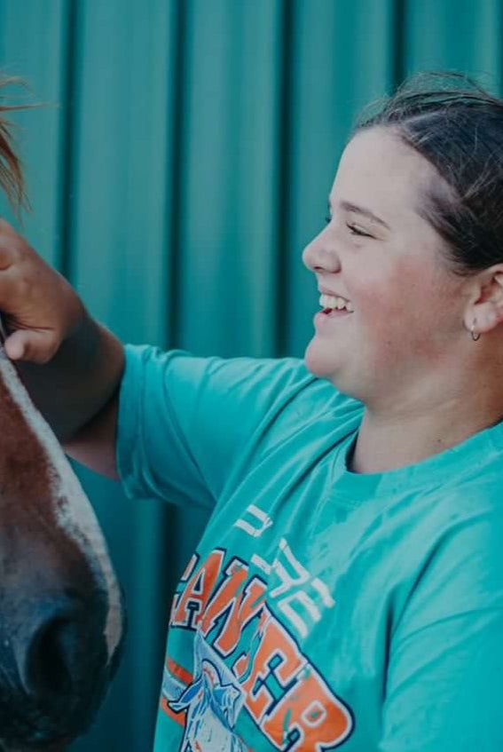 A young person with a ponytail wearing a green Youth PURE Sports Crop by Pure Canter Pty Ltd and high-waisted pants smiles while petting the face of a brown horse. The background is a green-striped wall.