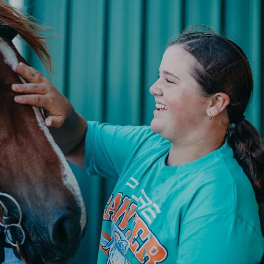 A young person with a ponytail wearing a green Youth PURE Sports Crop by Pure Canter Pty Ltd and high-waisted pants smiles while petting the face of a brown horse. The background is a green-striped wall.