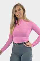 A woman with long blonde hair is smiling and posing against a plain white background. She is wearing a long-sleeved, fitted pink athletic top called the Motion Top Long - Pink from Pure Canter with a zippered collar made from moisture-wicking fabric and high-waisted grey pants.