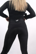 A person with long blonde hair is facing away from the camera, wearing a black long-sleeved top and Pure Canter's The Breeches - Black Leopard with a small white horse logo on the waistband. The breeches, made from performance fabric, have detailed stitching and zippered pockets on the back.