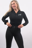 A person with long, blonde hair stands against a plain background, smiling slightly. They are wearing a black, long-sleeve zip-up top with a white logo on the chest and high-waisted The Breeches - Black Leopard from Pure Canter made from performance fabric. They have a black scrunchie on their left wrist.