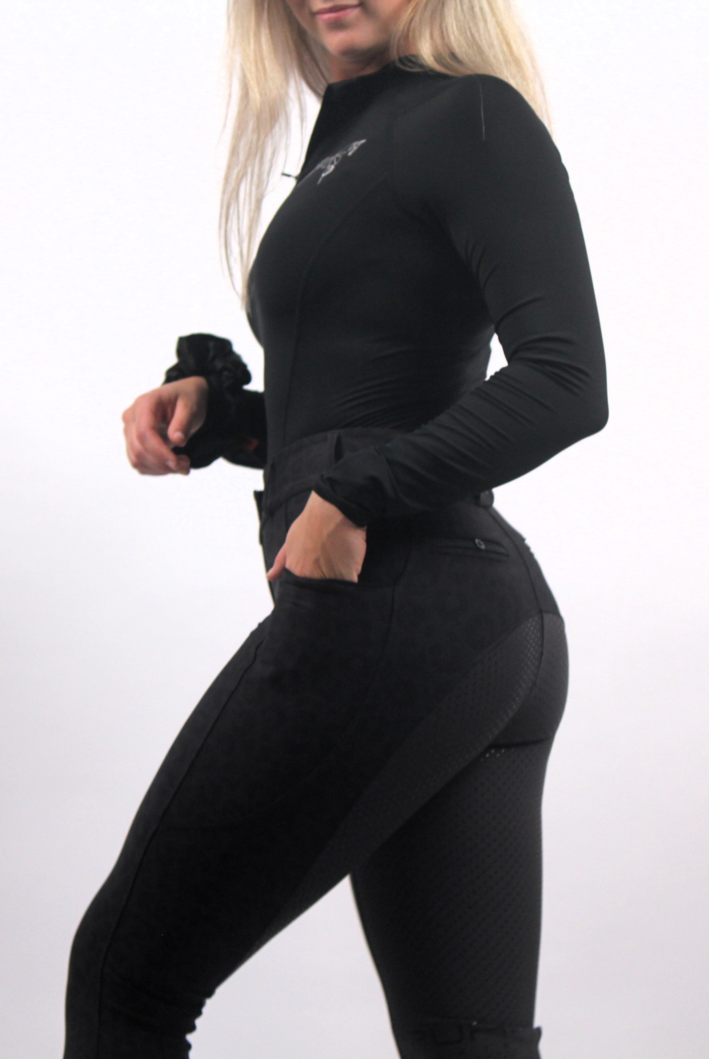 A person in black athletic wear stands on a plain background. They are wearing a long-sleeved black top and high-waisted The Breeches - Black Leopard by Pure Canter, made of performance fabric, with one hand in their pocket. Their face is partially out of the frame, and their long, light-colored hair is visible.