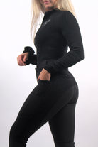 A person in black athletic wear stands on a plain background. They are wearing a long-sleeved black top and high-waisted The Breeches - Black Leopard by Pure Canter, made of performance fabric, with one hand in their pocket. Their face is partially out of the frame, and their long, light-colored hair is visible.